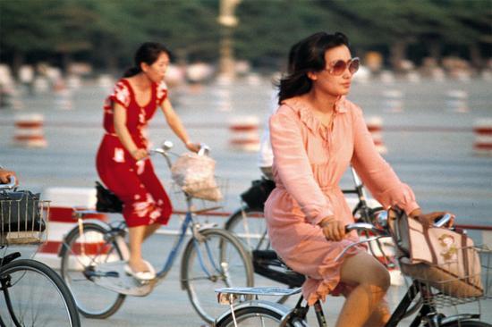 1985年夏,北京街头骑车的女青年。烫大波浪头、戴墨镜、穿粉色镶花边裙装,这位女性绝对是引领时尚的弄潮儿,她身后的红裙女士也毫不逊色。1984年,电影《街上流行红裙子》就反映了那一时期的社会风潮。摄影|李晓斌