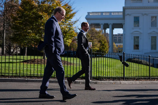 凯文・哈塞特双手插兜行走，身旁一名男子手持文件，背景可见白宫、绿色草坪和树木。