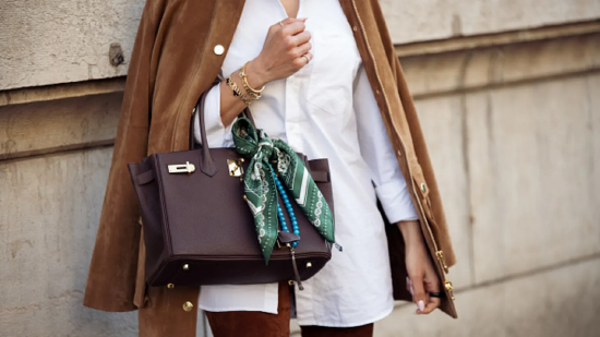 在 2025 年 3 月 8 日法国巴黎时装周 2025/2026 秋冬女装秀场外，一位嘉宾穿着白色衬衫、棕色裤子、棕色麂皮夹克，背着棕色爱马仕包，戴着绿白相间的丝绸围巾。