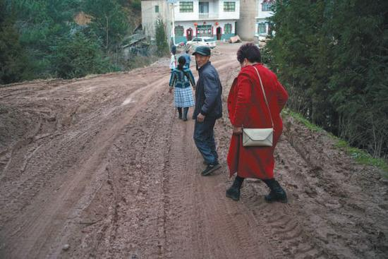 　▲村民从被拉矿的货车轧坏的路面小心翼翼走过，村民说，天晴一包灰，下雨一包浆，本来山清水秀的寨子现在污泥滂沱。　