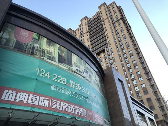 　　距离九州兴建棚改房一道之隔，为尚典国际商品房售楼中心，售楼部外部墙体张贴大幅海报，贴有“卖房送奔驰”“献给鹤岗万分之一城市英雄”等标语。