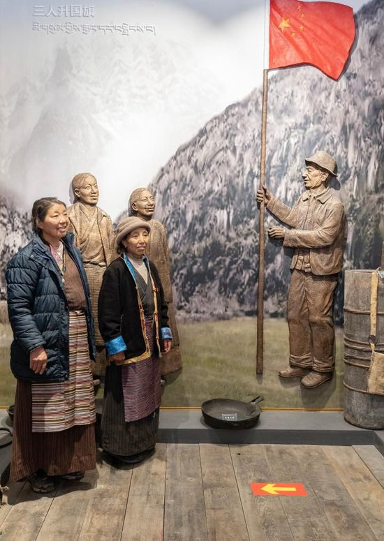 2023年6月10日，卓嘎（右）和央宗在桑杰曲巴故居中与描绘桑杰曲巴用自己缝制的国旗举行升旗仪式场景的壁画前合影。新华社记者 费茂华 摄