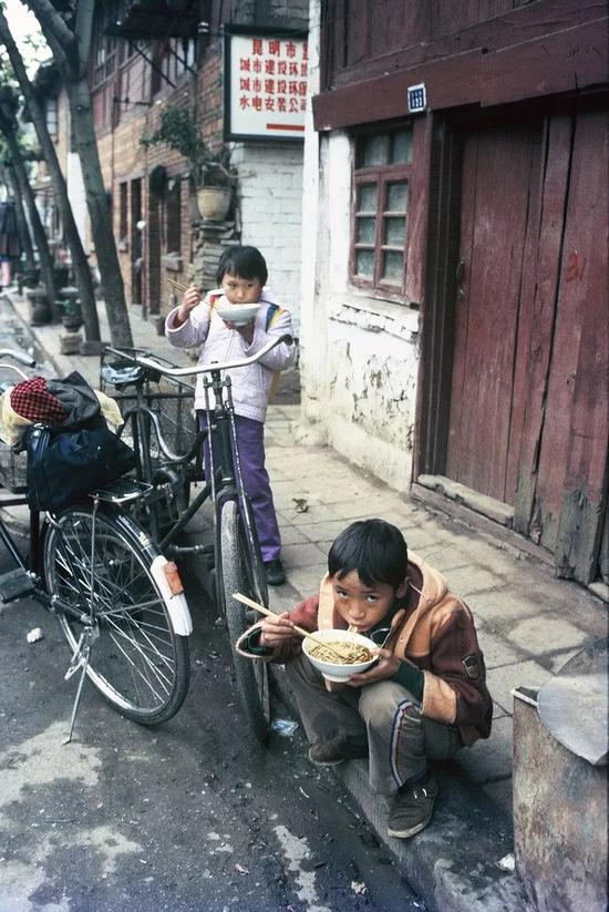 △ 1985年，昆明，在一条富有年代感的街道边，有两个小孩在吃面。