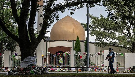 　　2019年3月22日，新西兰克赖斯特彻奇，武装警察在遇袭清真寺附近站岗，清真寺外摆满了哀悼的鲜花。图片来源：视觉中国