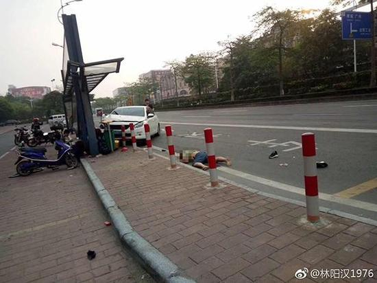 一辆白色小轿车的前身撞上公路边上的公交车站，一旁有人躺在路面上，疑似受伤。