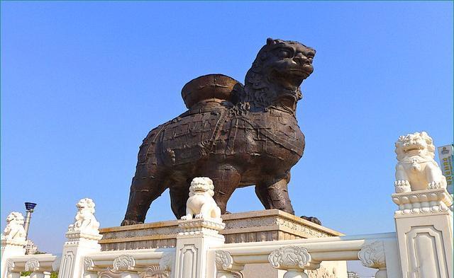 图片展示了一座雄伟的石狮雕塑，背后是晴朗的蓝天，狮子前有栏杆，上面还有几尊小型狮子雕像。