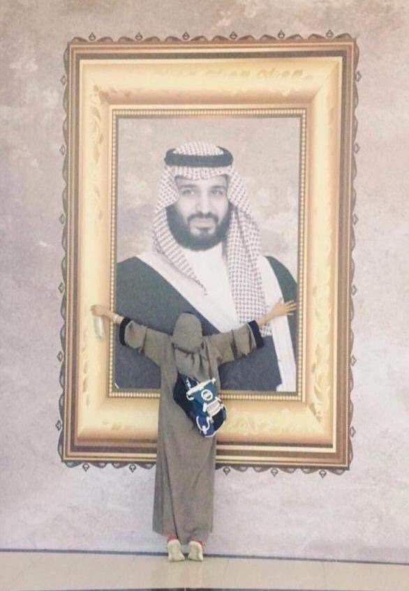 一位身穿灰色长袍、半裹头巾的女性拥抱巨幅王储画像。 采访对象供图