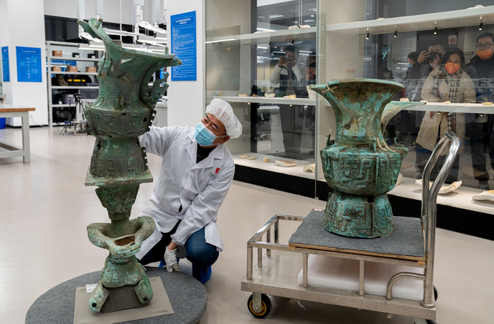 2021年12月23日，三星堆博物馆文物保护修复展示中心。郭汉中蹲下身子近距离研究今年发掘的重要文物顶尊跪坐人像。旁边为今年3月直播发掘的第一件重要文物青铜大口尊。观众透过玻璃观看国宝亮相。新京报记者 郭延冰 摄