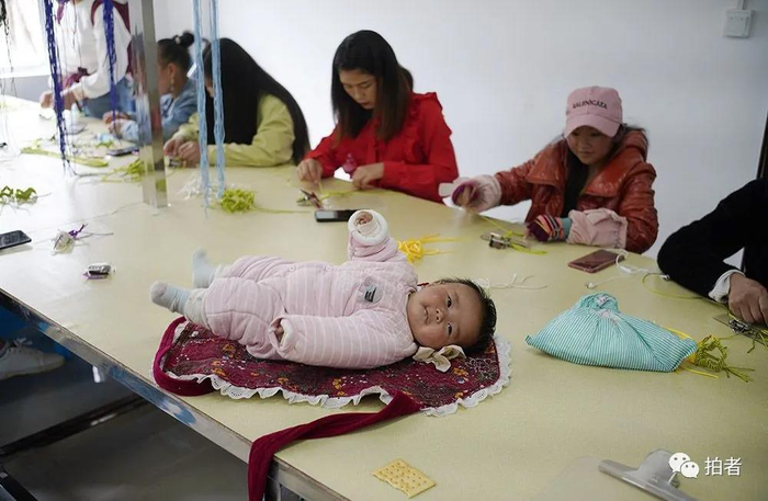 △ 2020年6月，贵州省威宁彝族回族苗族自治县五里岗易地扶贫搬迁安置点，欣荣家园扶贫车间，女工们带着孩子上班，有的就直接把婴儿放在工作台面上，让他睡在五颜六色的丝线边。摄影 / 新京报记者陈杰