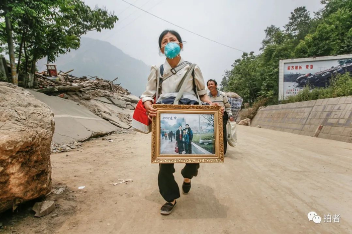 ▲2008年5月23日，老何的嫂子拿着何全寿一家三口的照片走出北川县曲山镇任家坪村。摄影 / 赵亢