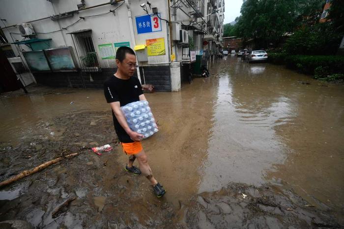 7月31日，北京门头沟，一男子抱着一箱饮用水走在积水的街道上。