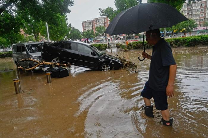 7月31日，北京门头沟，暴雨过后，一男子在被水淹没的街道上路过一辆受损车辆。