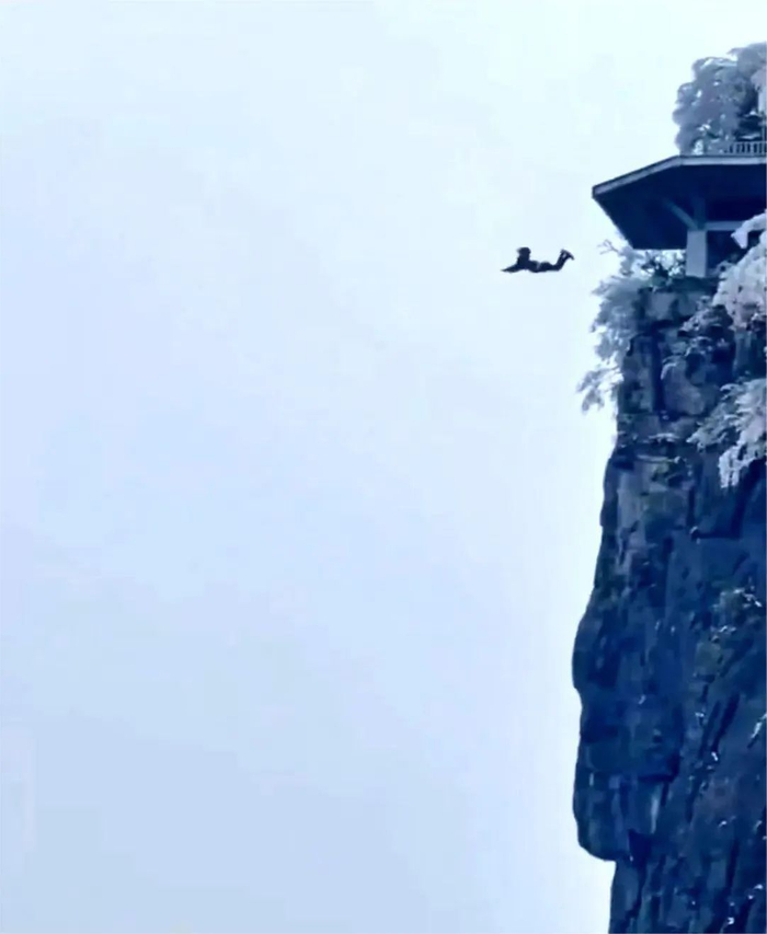 视频中,一处山体植被被白雪覆盖,一人站在接近山顶的观景台一跃而下.