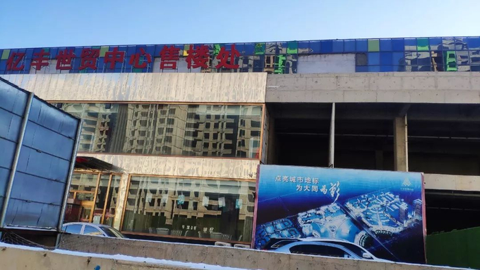 经过简单装修的售楼处与旁边空洞的主体结构形成鲜明对比，这场徘徊多年的烂尾困局至今未有终点。