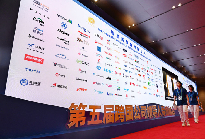 8月28日，两名志愿者从第五届跨国公司领导人青岛峰会跨国公司logo墙前经过。（李紫恒 摄）