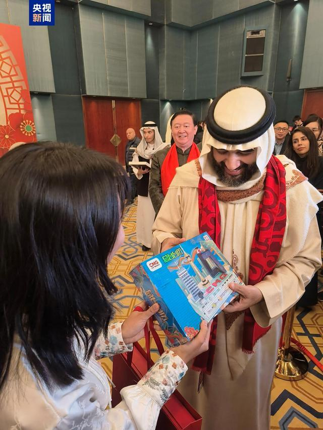 △沙特国家电竞协会主席班达尔亲王佩戴“巳巳如意”红围巾，并与总台记者共同展示总台文创产品