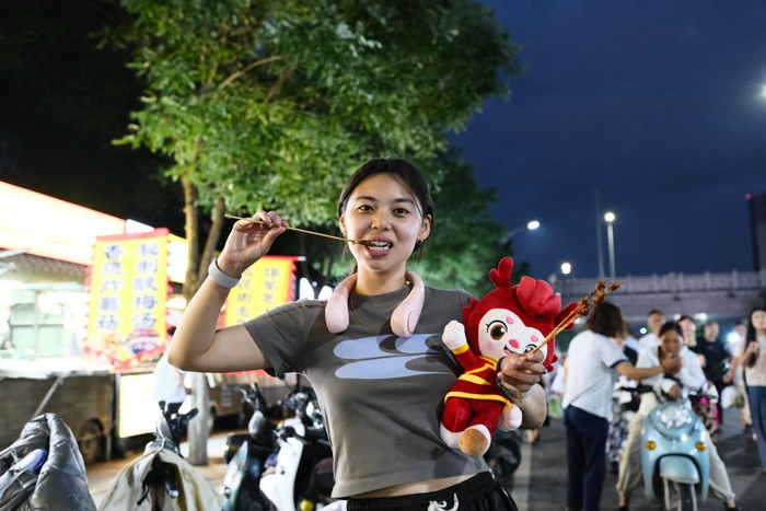 7月25日，在内蒙古赤峰市，抱着赛事吉祥物玩偶的游客在夜市品尝美食。 新华社记者 马金瑞 摄