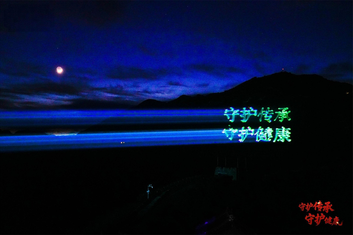 图为长城马道上“医圣小柴胡 健康全家福”的巨型发光字,以及远处山体映现活动主题“守护传承 守护健康”八个发光大字