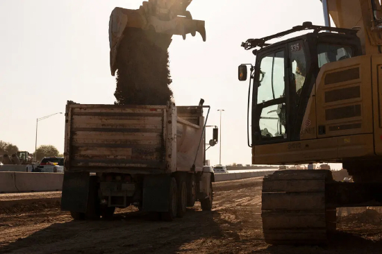 一台搭载 Bedrock Robotics 传感器的挖掘机正在向自卸卡车倾倒土方。Bedrock Robotics 表示，其产品最终可让建筑承包商实现 24 小时不间断作业。