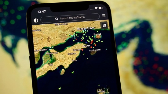 2026 年 4 月 8 日，法国克雷泰伊，一部智能手机屏幕显示 MarineTraffic 应用程序，背景为卫星视图，画面中霍尔木兹海峡附近密布大量船舶信标。当时美国与伊朗刚达成外交协议，重新开放这条战略水道供国际航运通行。